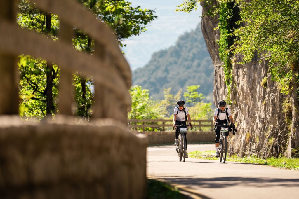 Two gravel riders on a road cut into the rock, with a lake and mountains behind