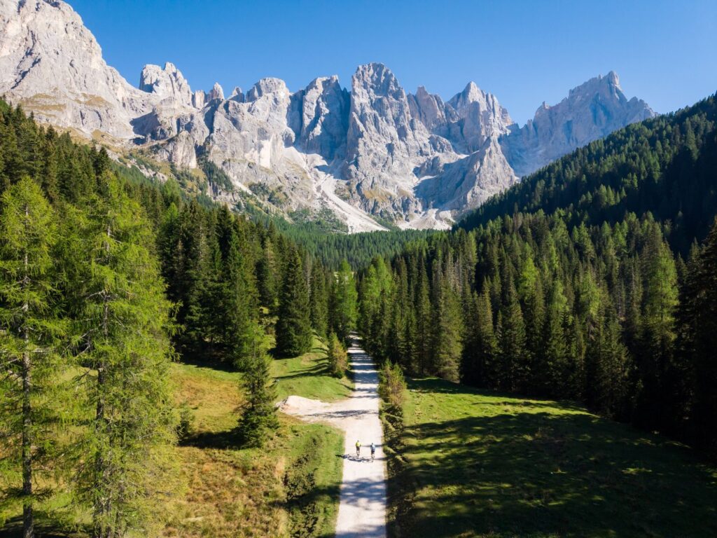 A gravel road crosses a valley of meadows closed by a wall of Dolomite spires