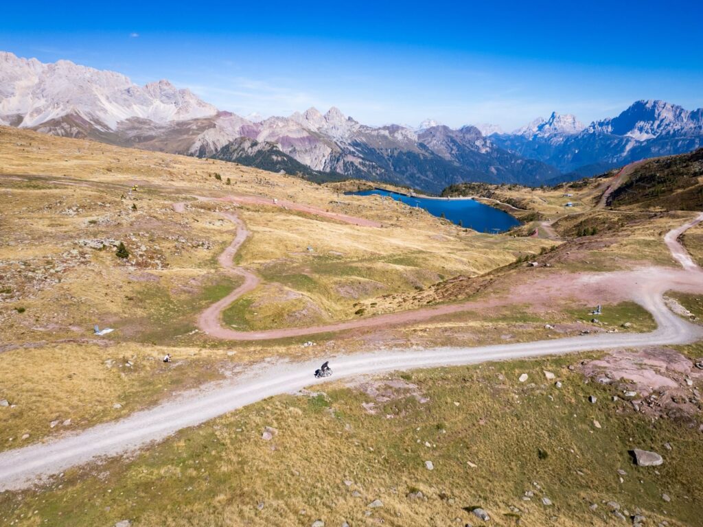 A gravel road winds across a high plateau toward a blue lake, Dolomites on the horizon