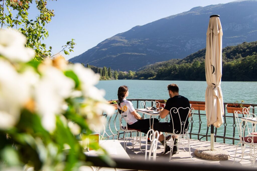 Two riders sit on a lakeside terrace among flowers, mountains in front