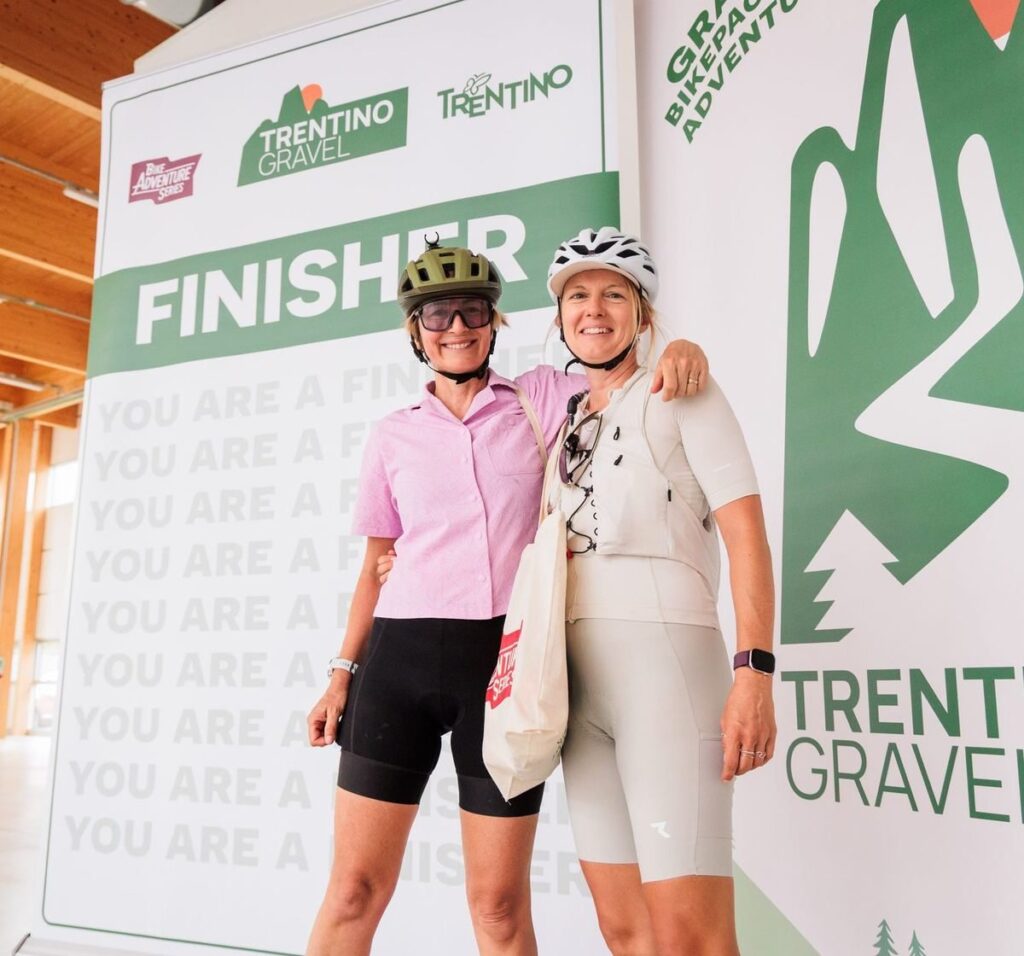 Two smiling finishers in front of the Trentino Gravel finisher wall